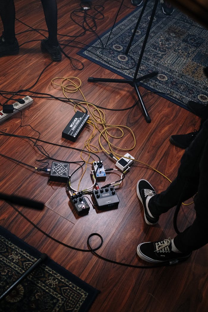 A view of audio equipment and pedals on the wooden floor of a music studio with musicians legs visible.